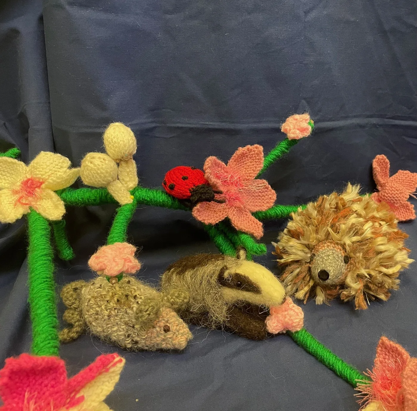 Knitted flowers on green stems with a knitted ladybird, badger, mouse and hedgehog laid out on a blue cloth