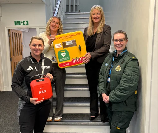 Four women on a staircase holding a red AED defibrillator and a yellow defibrillator cabinet, one of them in North West Ambulance Service uniform