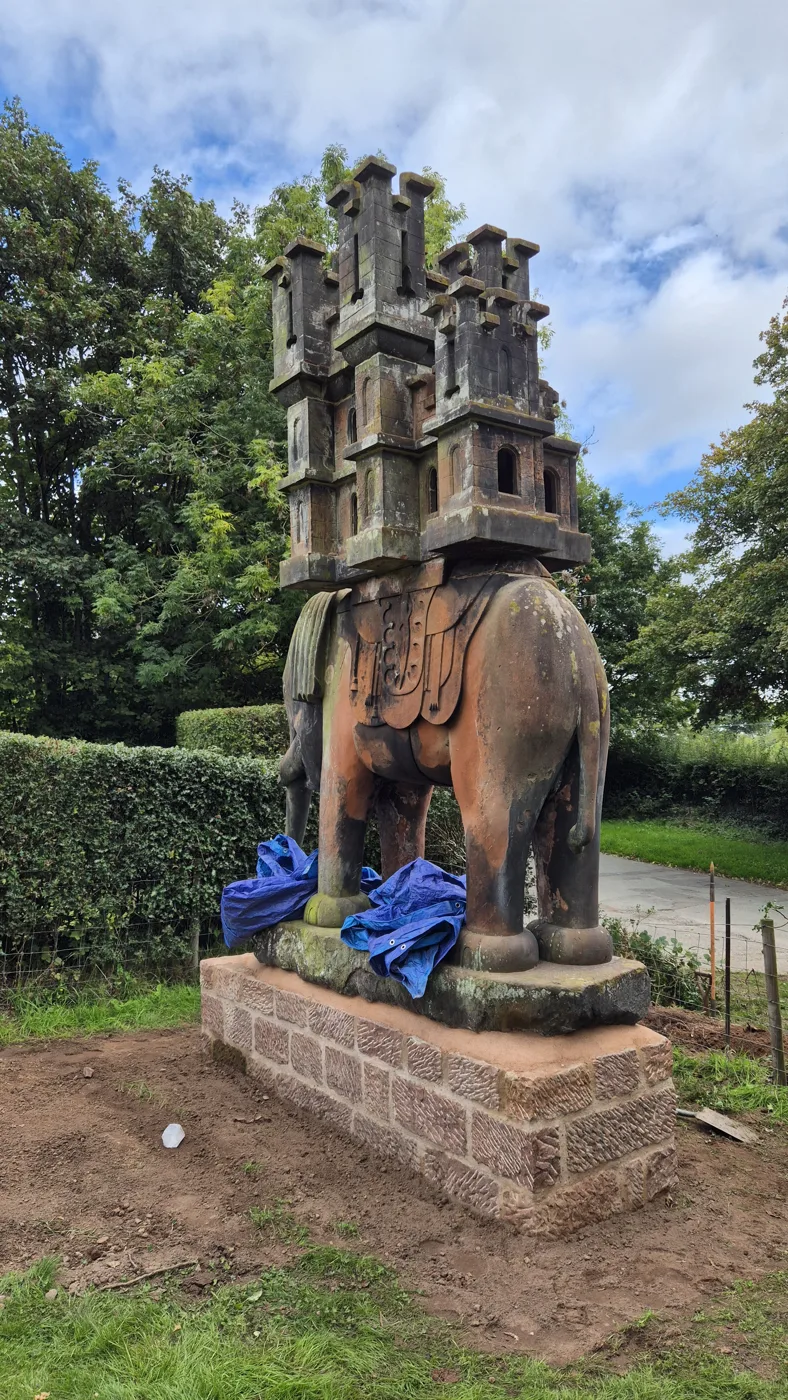 The sandstone elephant with its carved castle on its back, standing on a new dressed red sandstone plinth, with blue sheeting around its feet and bare earth around the base