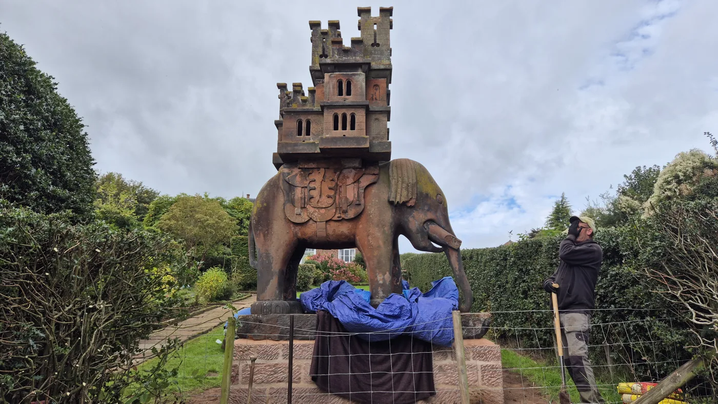 The elephant and castle on the new plinth, with blue sheeting and a dark cloth over the front of the base, behind a wire fence, and a man with a spade beside it