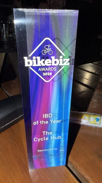 A glass BikeBiz Awards 2026 trophy reading IBD of the Year, The Cycle Hub, sponsored by V12 Retail Finance, standing on a wooden table